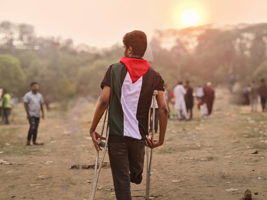 A young man wrapped in a Palestinian flag walks with crutches through a field in Bangladesh during the day.